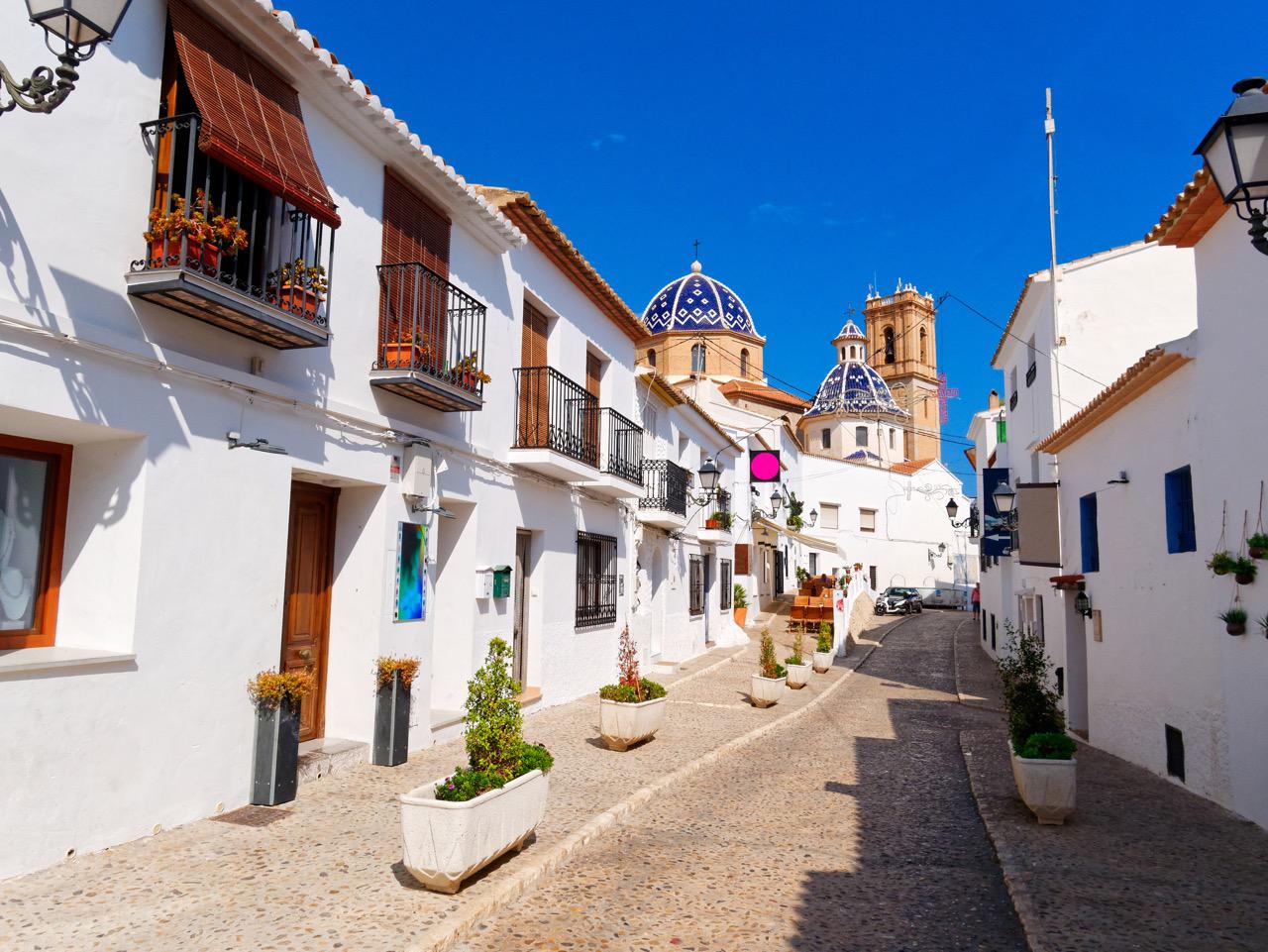 Narrow street lined with white buidlings in Altea, and the iconic blue-domed church Nuestra Señora del Consuelo in the distance